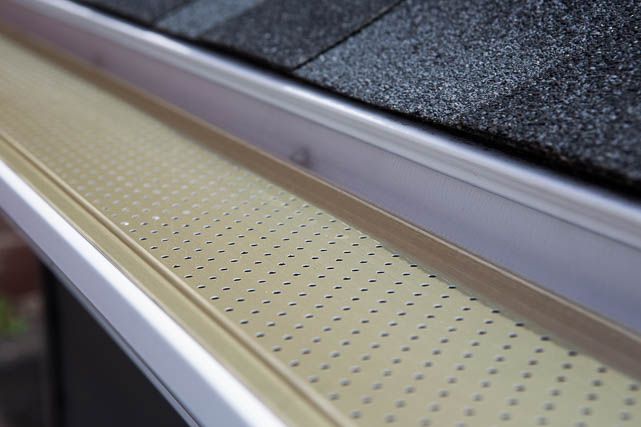 Close-up of a home's eavestrough installations, featuring a perforated metal cover over the gutter. The roof shingles are visible in the top right corner of the image. The metal cover is designed to prevent debris from clogging the gutter while allowing water to flow through.