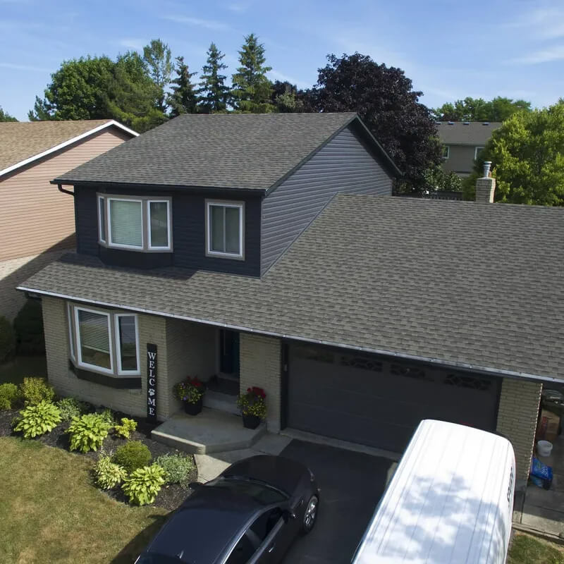 A two-story house with dark gray siding and a shingled roof, featuring a "Welcome" sign near the front entrance. The driveway has a black car and a white vehicle parked on it. The house is surrounded by neatly trimmed bushes and trees.