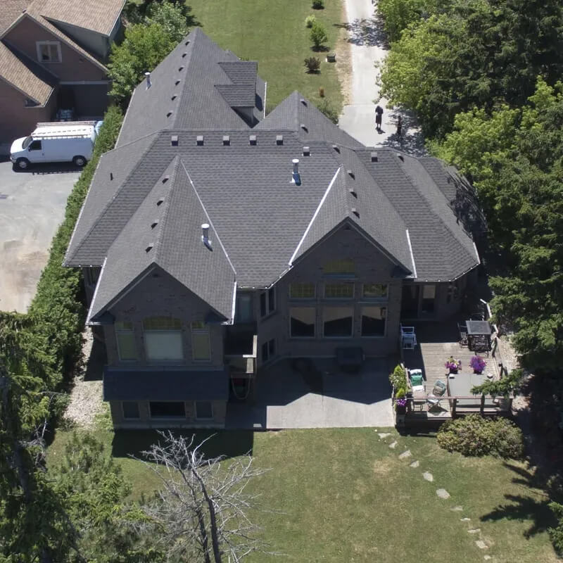 Aerial view of a large, two-story house with multiple gabled roofs, surrounded by trees. The house has a driveway leading to a road at the top of the image. A white van is parked to the left, and a small garden with outdoor seating is visible on the right.