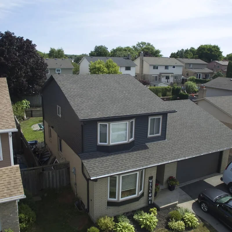 Aerial view of a suburban home with grey roofing, dark blue siding on the upper level, and light brick on the lower level. The front yard features a "Welcome" sign and manicured landscaping. Neighboring houses can be seen in the background.