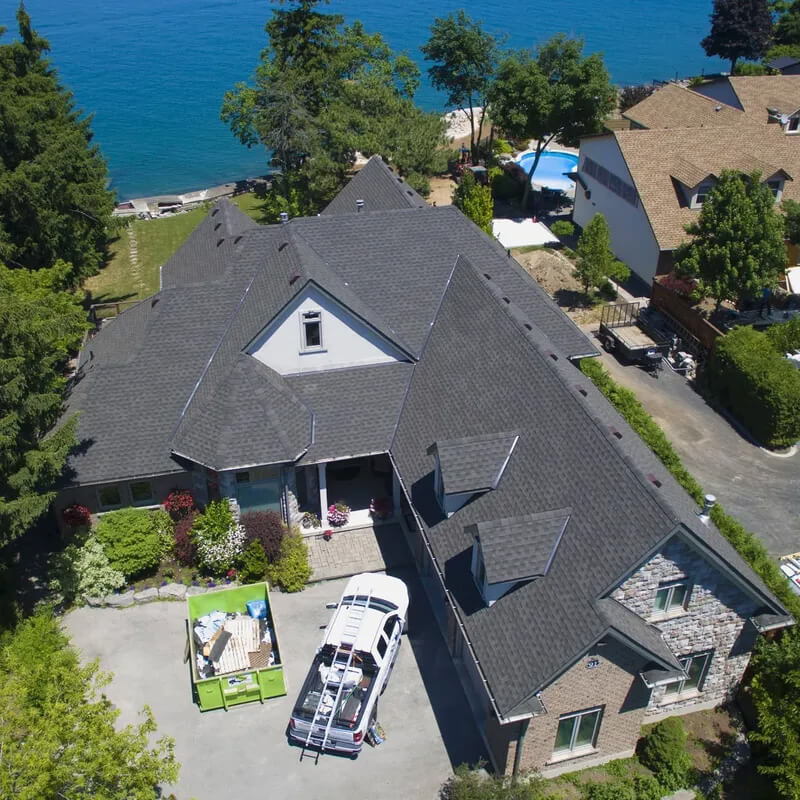 Aerial view of a large house with a dark gray roof located near a body of water. The property has surrounding trees and a driveway with a truck and container box. Other houses are visible adjacent to the property.