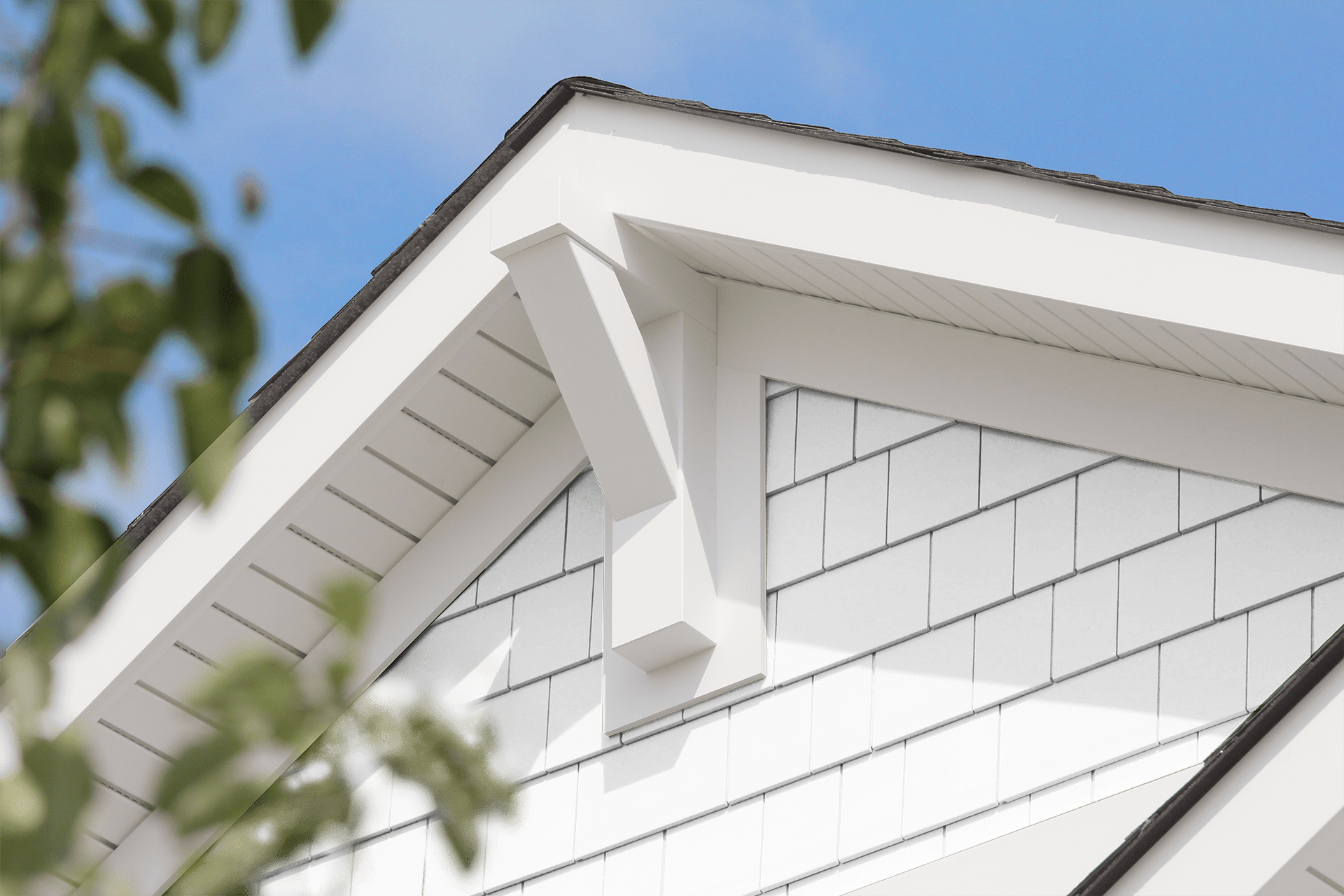 Close-up view of the roof of a white house with black shingles and a decorative white roof bracket. The pitched roof has dark shingles, and a blue sky is visible in the background. A leafy branch is partially visible on the left side of the image, showcasing Siding Services Hamilton's detailed work.