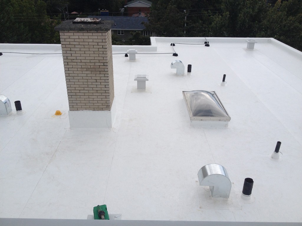 A flat white rooftop, installed by expert commercial flat roofing installers, features several vents, a brick chimney, and a skylight. Trees and rooftops of nearby houses are visible in the background under an overcast sky.