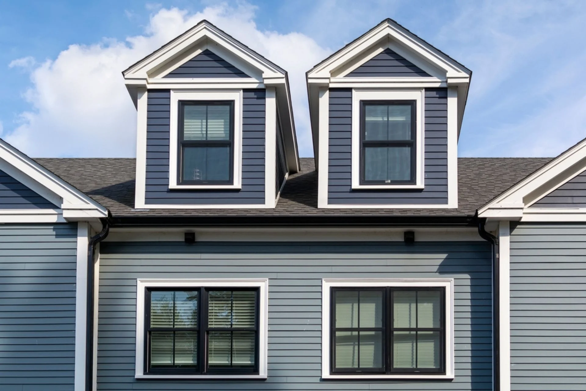 A two-story house with blue siding and white trim, featuring two dormer windows on the upper level and two larger windows on the main level. The sky is clear with a few clouds, showcasing quality work that could be matched by Siding Services Hamilton.