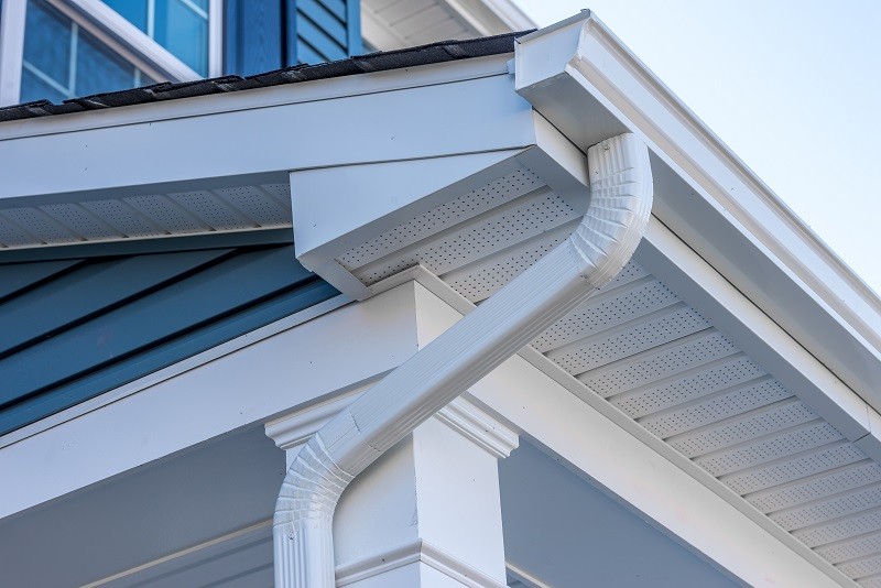 Close-up view of a house's gutter system, featuring a white downspout and soffits under the eaves. The house exterior, painted blue with white trim, highlights part of the roof and sky in the background. This perfect example of eavestrough installations shows attention to detail and functionality.