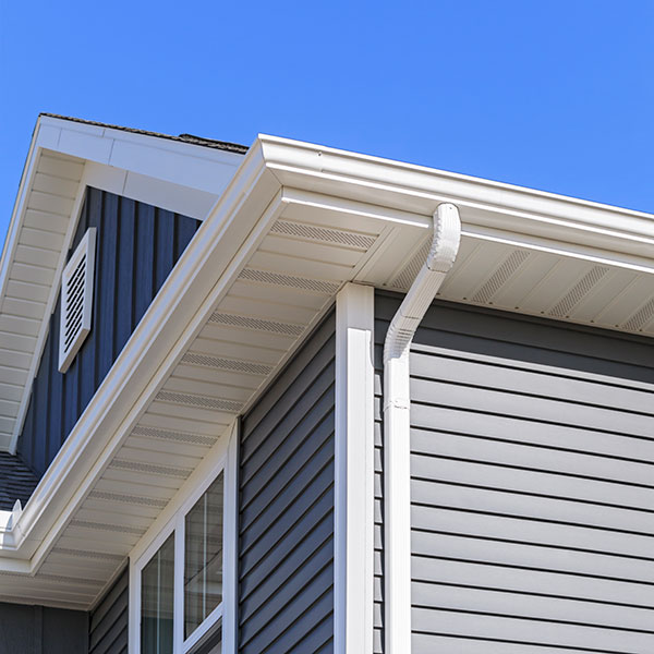 A close-up view of a house roof corner featuring a grey exterior with white trim. The image highlights a white eavestrough installation attached to the roofline. The sky is clear and blue, providing a contrast to the house's color scheme.