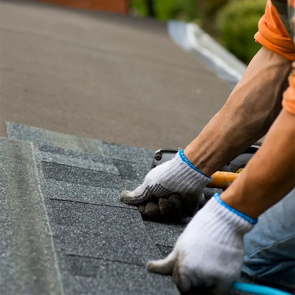 A close-up of a person installing roof shingles during a Roof Repair Hamilton project. The person is wearing white gloves and working with tools on a rooftop, focusing on placing the shingles evenly. The background is slightly blurred, emphasizing the roofing task at hand.