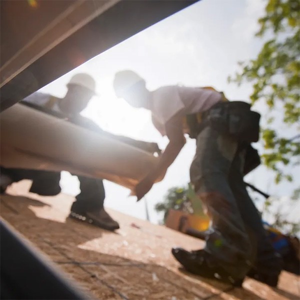 Two construction workers are seen from a low angle on a sunny day, handling a large board or pipe on a rooftop. They wear hard hats and safety vests. The photo captures their silhouettes with bright sunlight in the background, partially obscuring details—a perfect day for Roofing Services Hamilton.