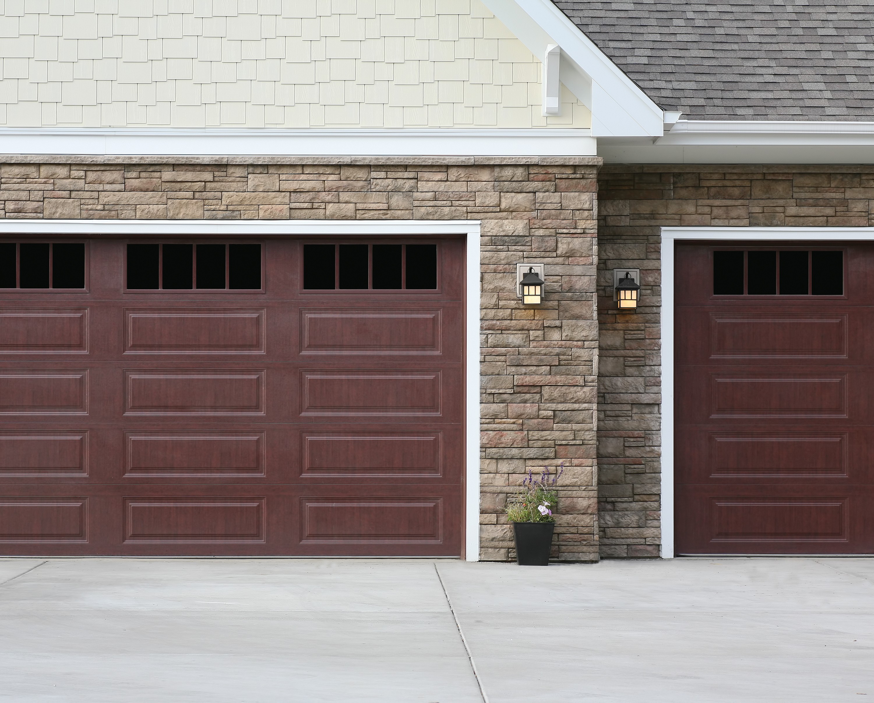 Two closed garage doors with a wooden finish are set against a stone wall facade, showcasing the quality you'd expect from premium siding services in Hamilton. Each door features a row of small square windows at the top. A black outdoor wall lantern is mounted between the doors, and a potted plant sits nearby.