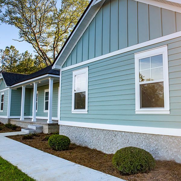 A modern single-story house with light blue siding, white trim, and a gabled roof. The home features a small front porch with white railings and columns and two square windows on the visible side. Landscaping includes a concrete walkway and trimmed bushes—quality work by Siding Services Hamilton.