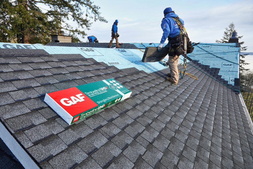 Roofers from Gateway Exteriors are installing slate-colored asphalt shingles on a residential roof. Using GAF-branded materials, one worker is lifting shingles while another secures them in place. A large package of GAF shingles sits on the roof, ready for use.