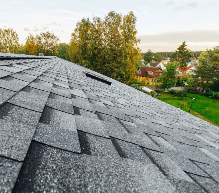 View from the edge of a shingled roof, looking over a lush green neighborhood with houses scattered among trees and a slightly clouded sky overhead. The focus is on the textured gray shingles in the forefront, highlighting the quality craftsmanship synonymous with Gateway Exteriors.