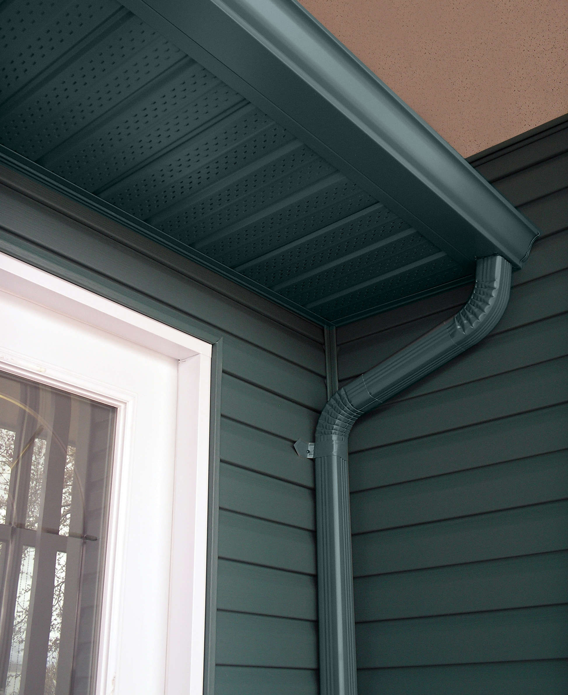 A corner of a house in Hamilton featuring green siding, a white door with a window, and a green gutter system including a downspout. The gutter is affixed to a perforated soffit under the roof eave. The scene is brightly lit, showcasing clean and well-maintained exterior elements, thanks to regular roof maintenance.