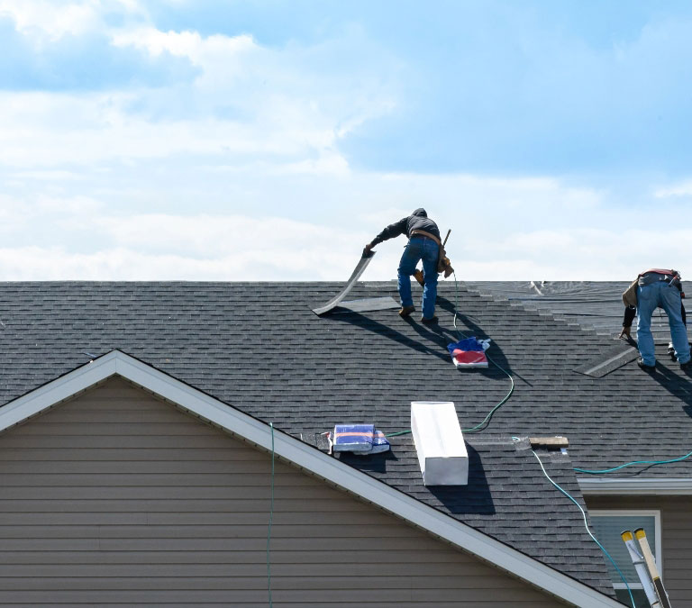 Two workers wearing safety gear are on the sloped roof of a house, installing new shingles. Tools and materials, including a ladder, a coil of cable, and packages of shingles from Gateway Exteriors, are scattered around. The sky is partly cloudy in the background.