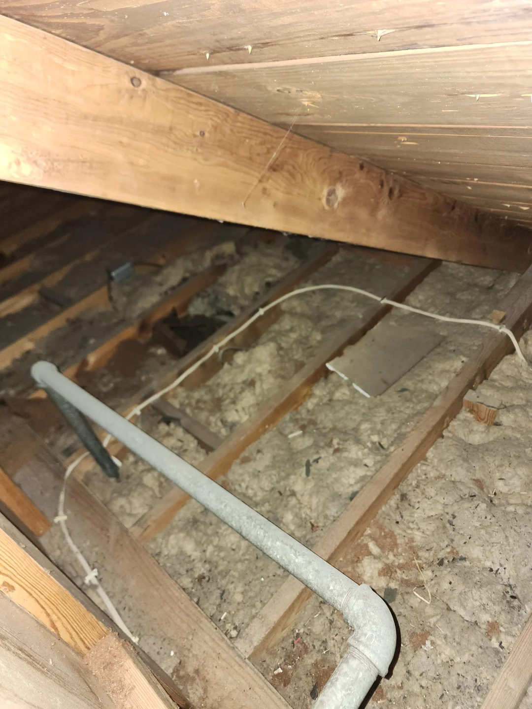 A dimly lit attic with wooden beams and insulation material between the floor joists. There are exposed pipes running through the space and some electrical wires visible against the wooden structure, highlighting the need for roof maintenance in Hamilton.