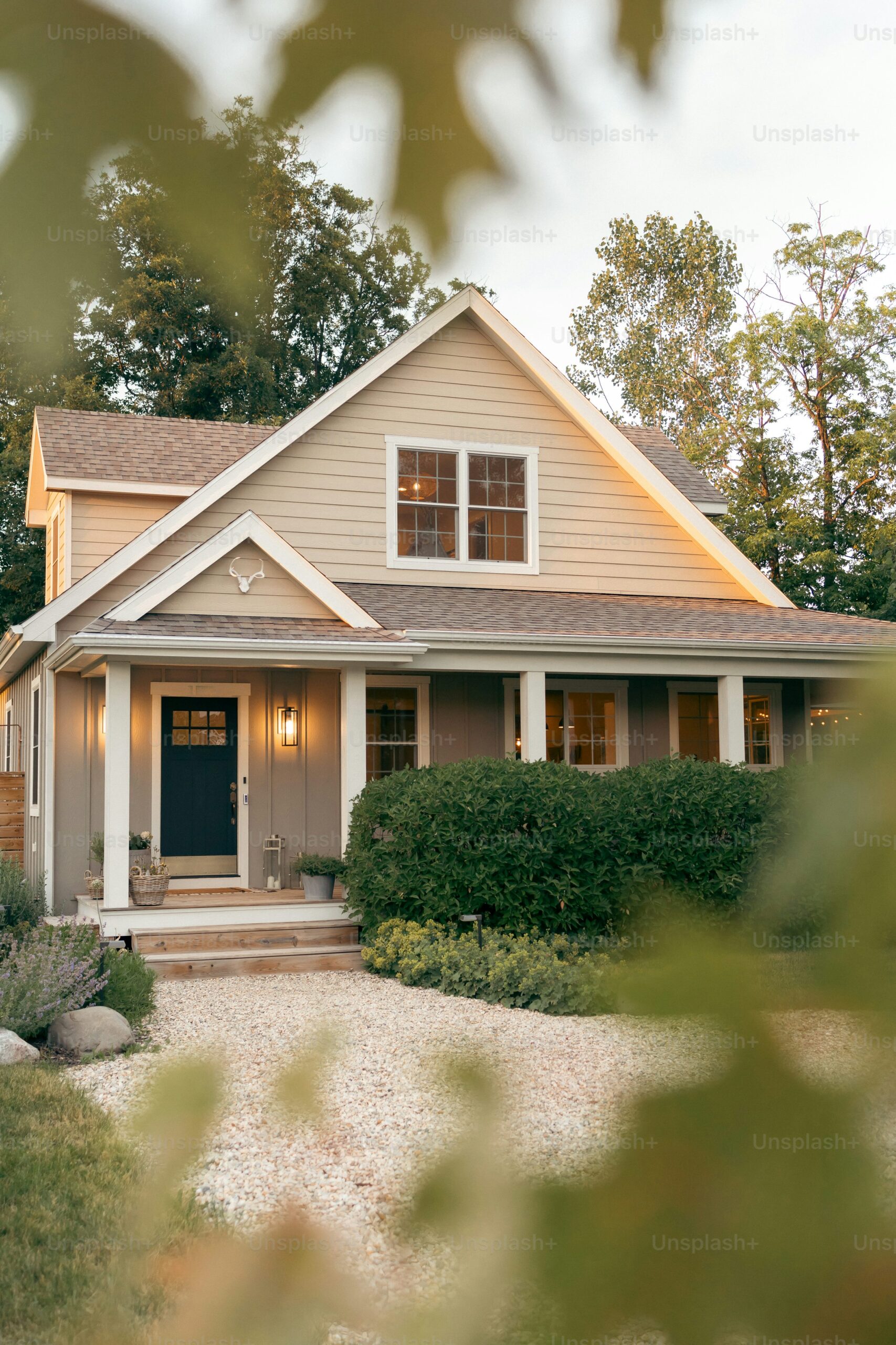 A charming two-story house with a beige exterior, black front door, and a wide front porch adorned with white railings and columns. The yard features a lush green hedge and a gravel pathway leading to the entrance, framed by leafy branches in the foreground. Perfect for those seeking roof maintenance in Hamilton.