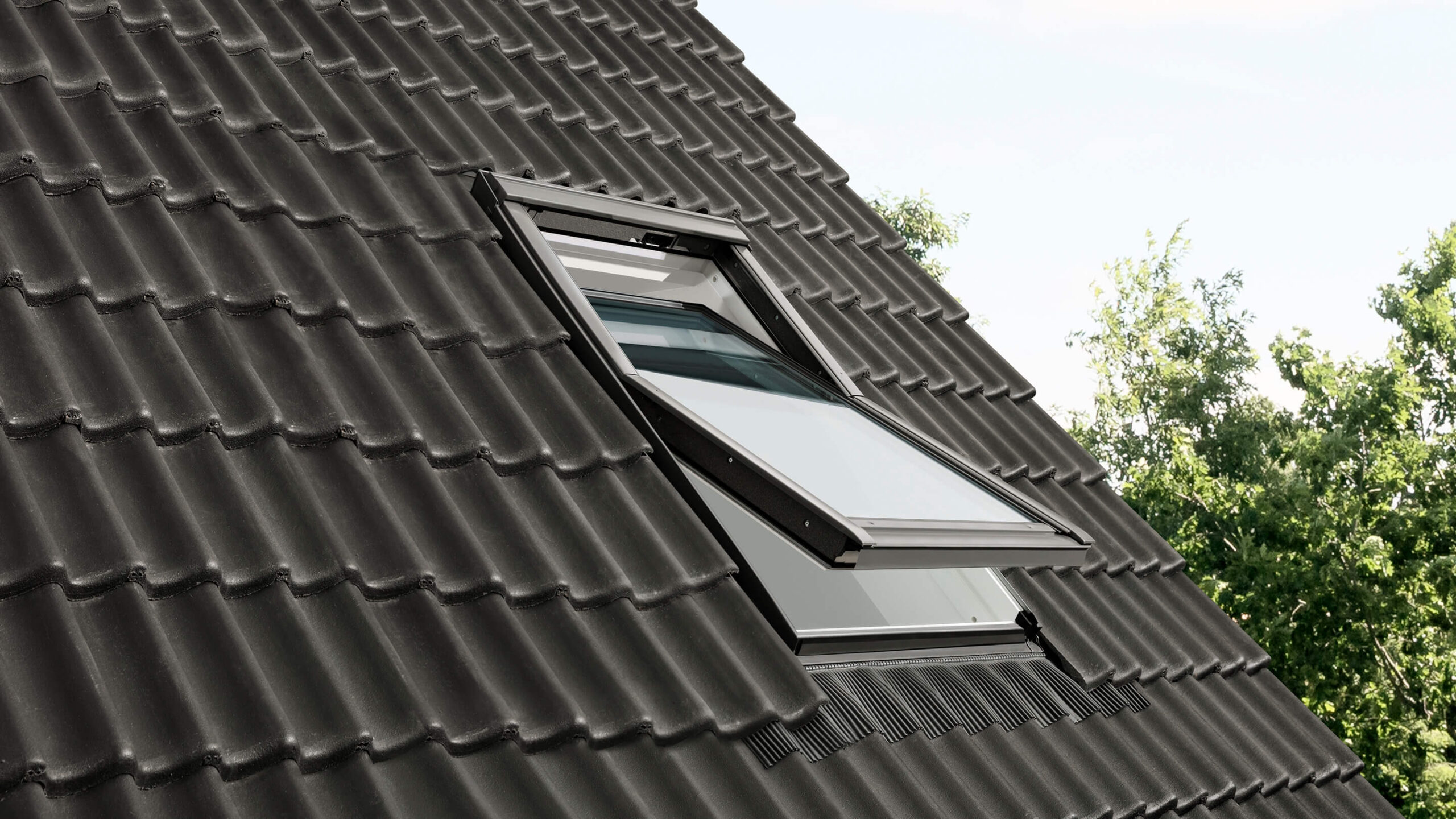 A close-up of a dark grey, tiled rooftop with an open skylight window. Gateway Exteriors showcases the window slightly raised, revealing a bright sky and green foliage in the background.